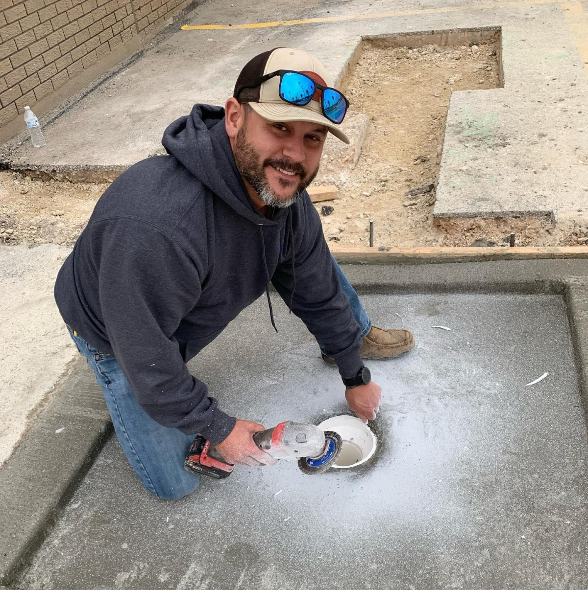 A plumber in a hoodie and cap drilling into a concrete surface with a power tool, preparing for plumbing installation.