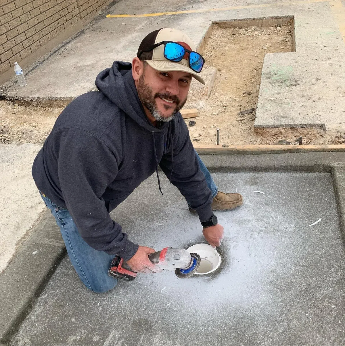 Yard Leak Detection in San Antonio 3 A plumber in a hoodie and cap drilling into a concrete surface with a power tool, preparing for plumbing installation.