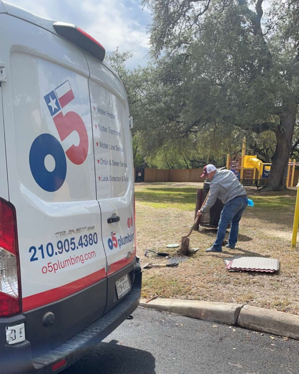 A plumber performing outdoor maintenance near a park, standing beside a branded plumbing service van.