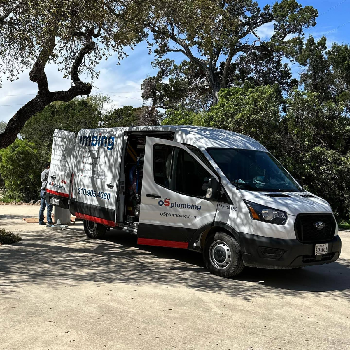 A plumbing service van with company branding parked on a driveway surrounded by trees.